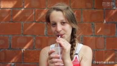 漂亮的小女孩坐在砖墙前，用吸管从塑料水杯里喝水